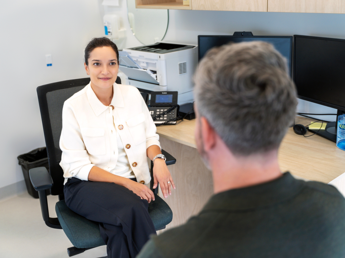 A female professional or doctor in a white jacket sits at her desk, smiling and listening attentively during a consultation with a male client or patient seen from behind.