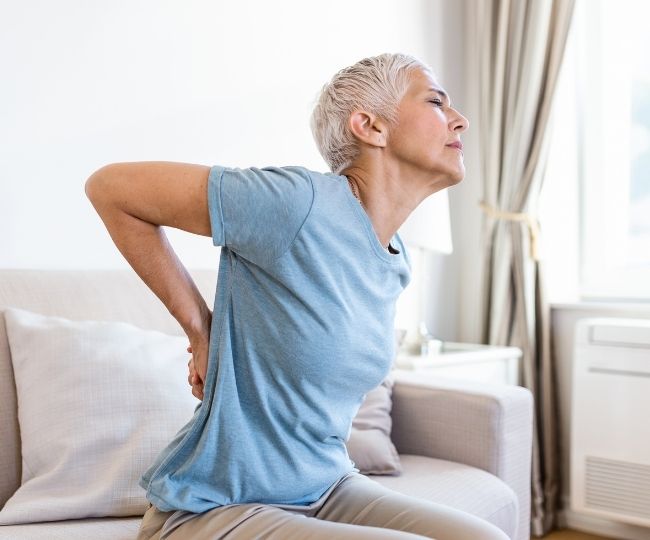 Older woman sitting indoors and holding her lower back with a pained expression.